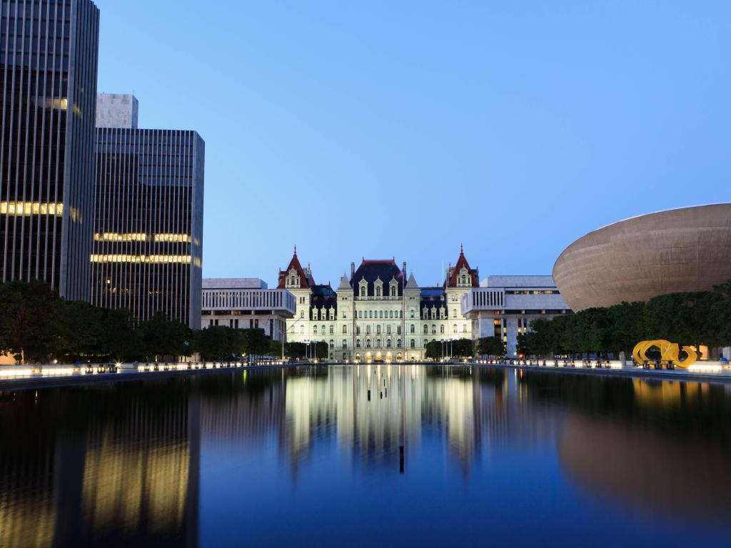 Historic white State Capitol building viewed across still lake water with high rise buildings to one side and a modern building to the other