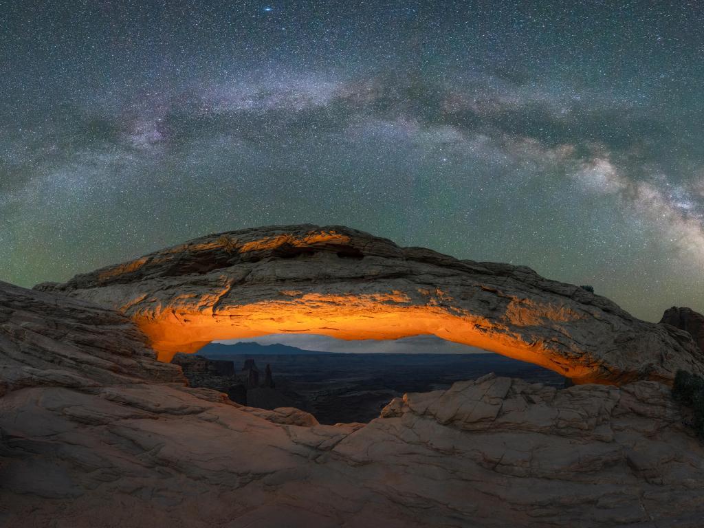 Night time view of Milky Way Galaxy panorama over a lit Mesa Arch in Canyonlands National Park, Utah