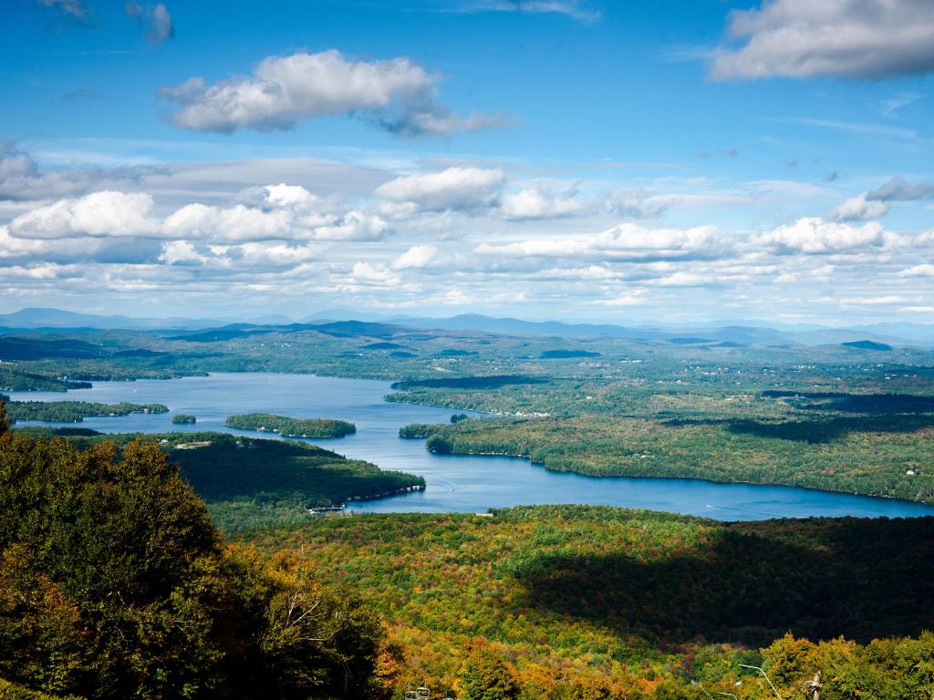 Sunapee Lake, New Hampshire, USA with an aerial view of the lake on a cloudy but sunny day, surrounded by greenery and hills in the distance.