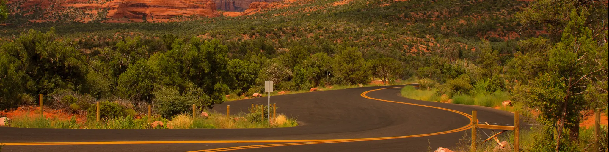 An image of an empty road in Sedona, Arizona during sunset with trees and a view of mountains.