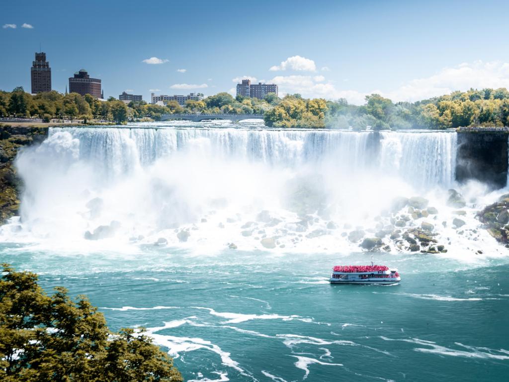 Niagara Falls, New York, USA taken on a sunny day with a boat in the water and the falls in the distance with trees and buildings in the background.