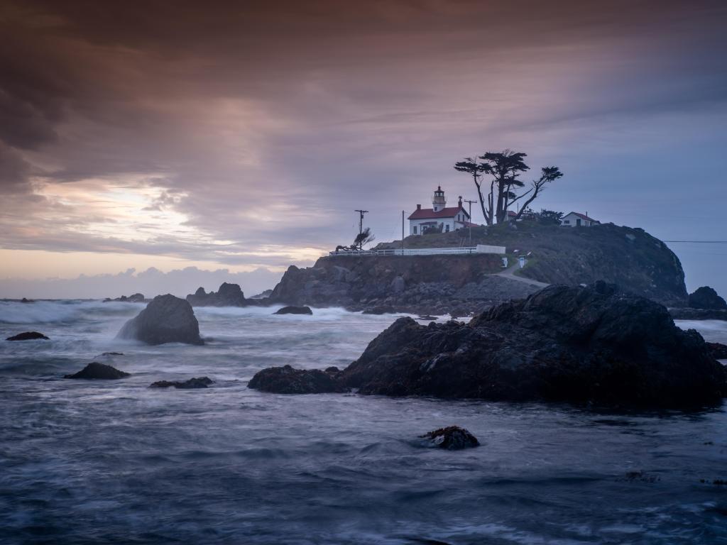 Choppy sea breaks on rocks in the water around the lighthouse up on a small island, with grey, stormy sky