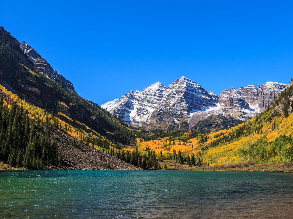 White River National Forest, Colorado, USA with a view of the Maroon Bells and a lake in the foreground, trees and snow-capped mountain in the distance against a blue sky.