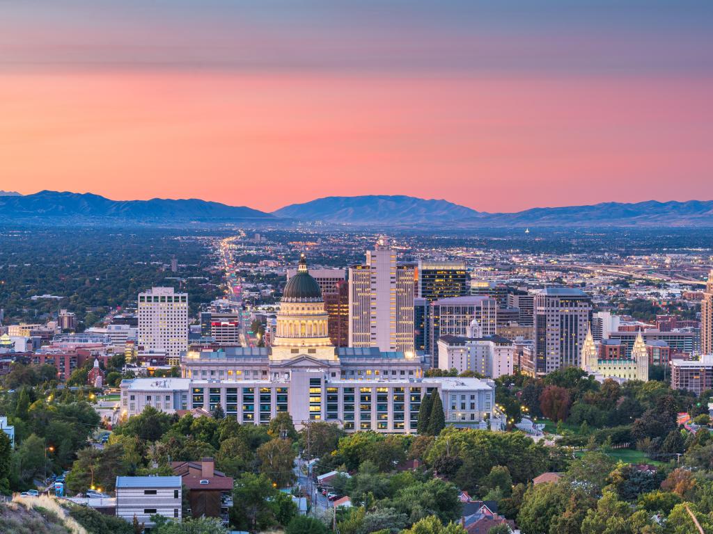 Salt Lake City, Utah, USA with a view of the downtown city skyline at dusk and mountains in the distance against a pink sky.