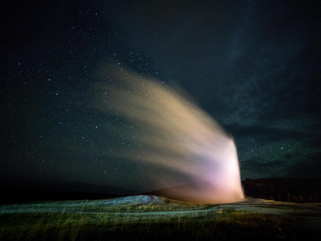 Hot steam erupts in a fountain from a geyser, brightly lit by moonlight in a dark sky filled with stars