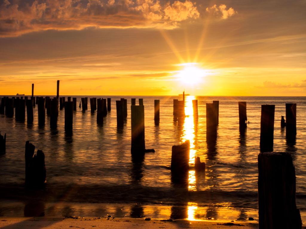 A ship crosses under the sun making a cross with the silhouetted posts from an old ferry dock during a golden sunset