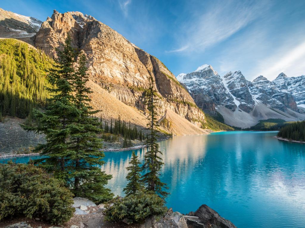 Lake Moraine, Banff National Park, Canada taken on a sunny day with trees in the foreground, the blue lake and mountains in the distance with snow on the very tops.