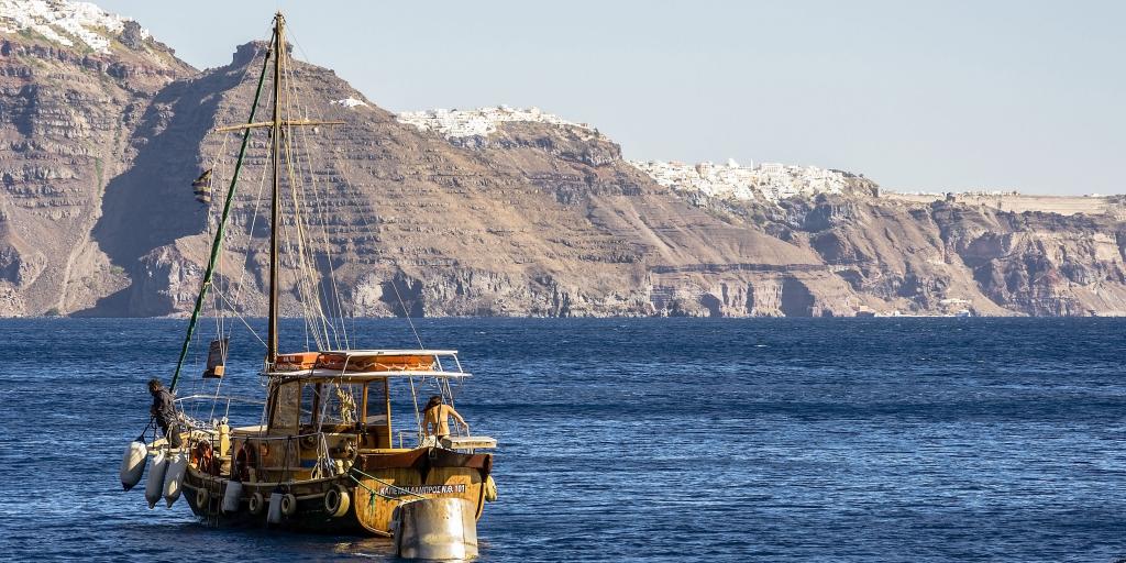 A wooden ship sails in the deep blue water in front of the red cliffs and white villages of Santorini