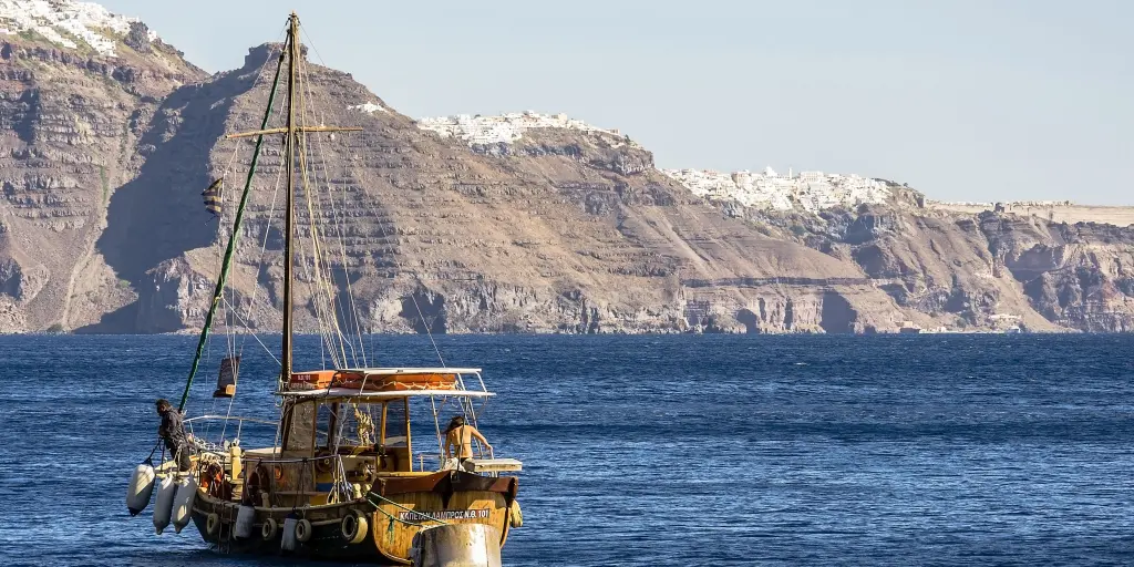 A wooden ship sails in the deep blue water in front of the red cliffs and white villages of Santorini