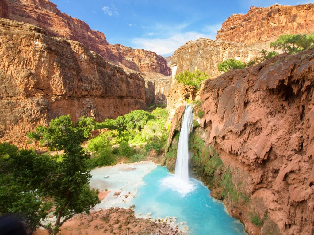 Havasu Falls, Grand Canyon National Park, USA taken on a sunny day with the waterfall surrounded by red rock cliffs, trees and taken on a sunny day.