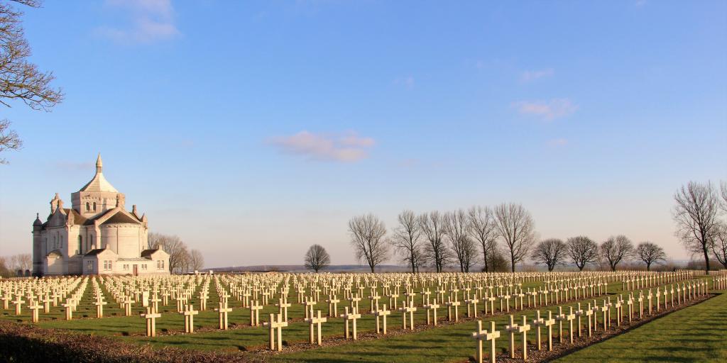 The white chapel at Notre Dame de Lorette Cemetery, France, with rows and rows of graves with white crosses in front and bare winter trees behind