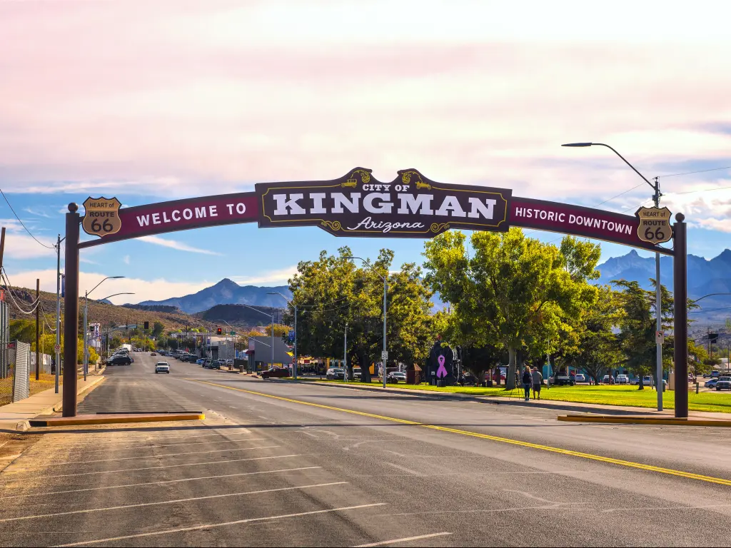 An arches street sign located on historic route 66 Kingman, Arizona with cars driving along the road, people walking in the sidewalk under the shade of trees, and the view of the mountain from the distance