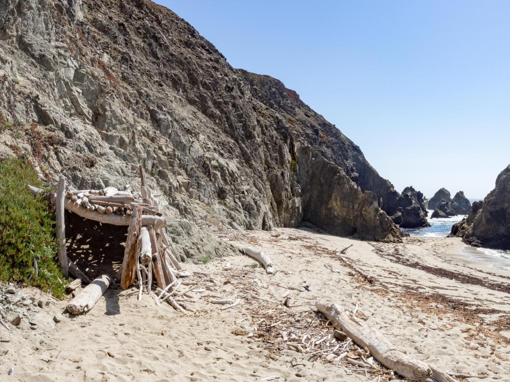 Bodega Head, California on a sunny day with a driftwood shelter on beach in the foreground and rocky cliffs leading into the distance. 