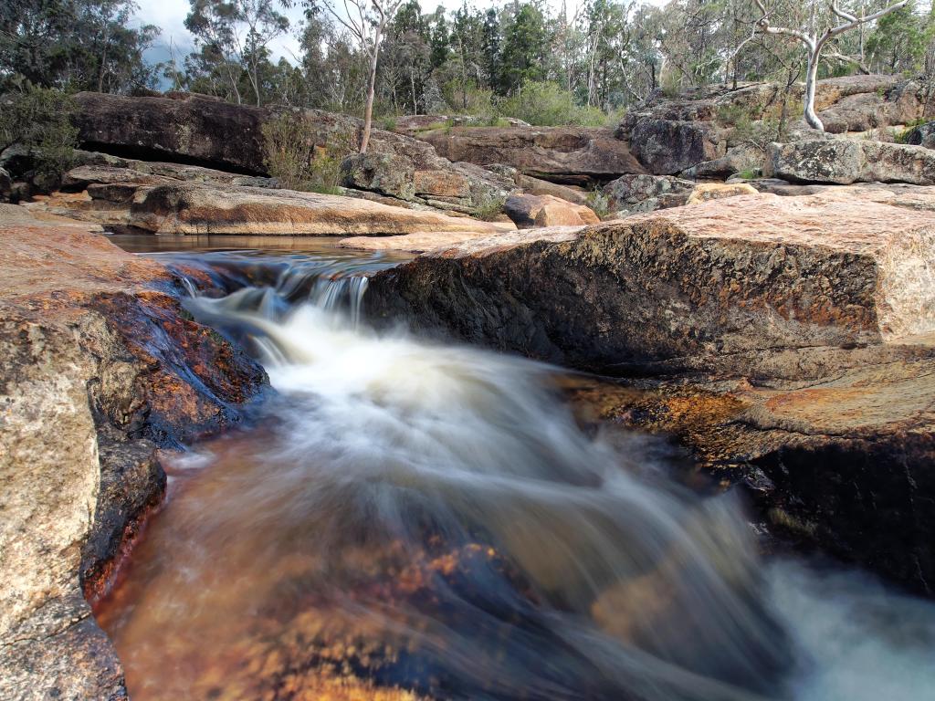 River cascading down small waterfall across flat rocky riverbanks at Woolshed Falls, Beechworth, Victoria, Australia