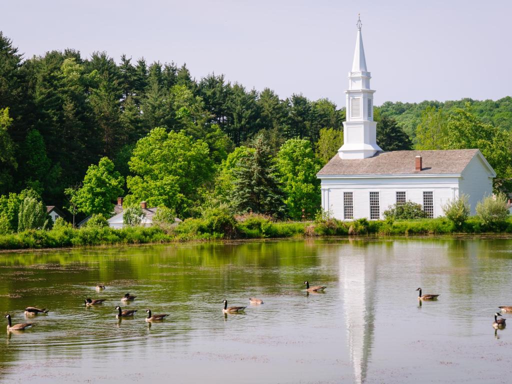 Small white church beside a lake with ducks swimming, surrounded by green trees