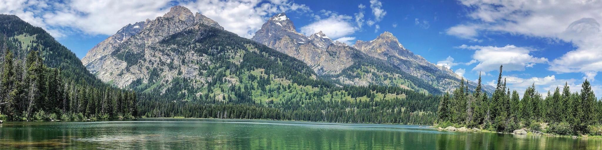 Grand Teton National Park, Wyoming with a reflection of the forest and mountains in the lake in the foreground on a cloudy blue sky.