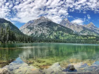 Grand Teton National Park, Wyoming with a reflection of the forest and mountains in the lake in the foreground on a cloudy blue sky.