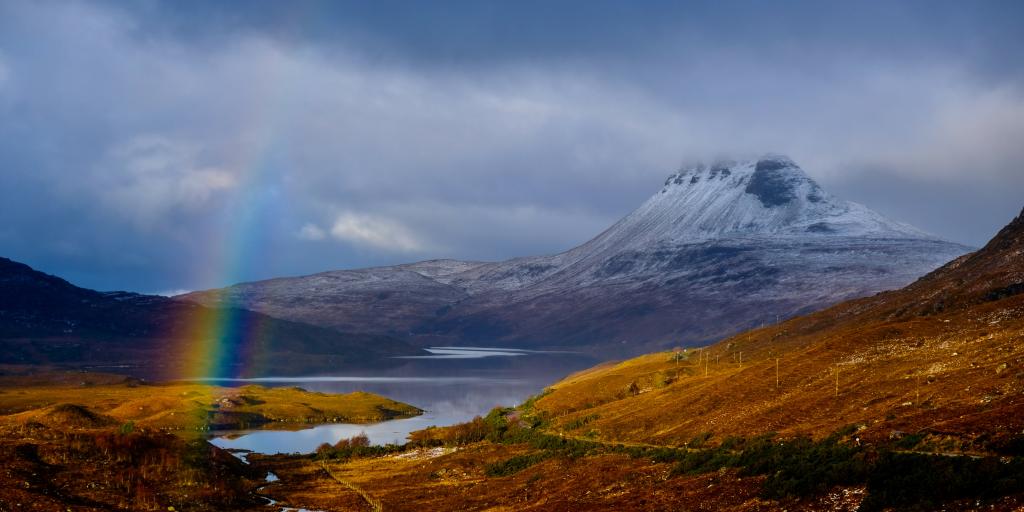 Stac Pollaidh Mountain, Scotland, coated in snow on a cloudy day, with a rainbow and a loch in front