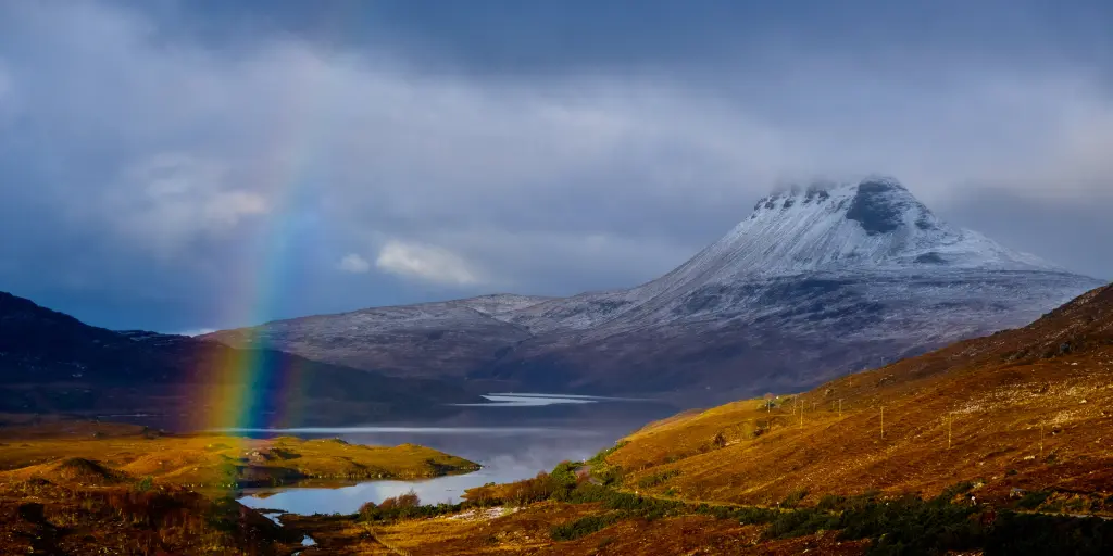 Stac Pollaidh Mountain, Scotland, coated in snow on a cloudy day, with a rainbow and a loch in front