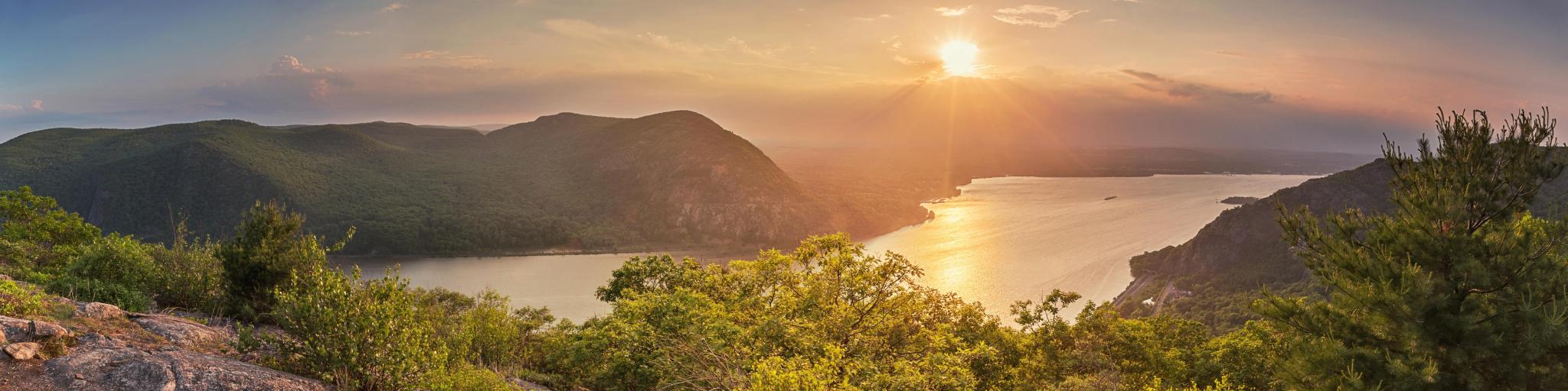 View over wide Hudson River with green trees covering the steep river sides and a bright setting sun
