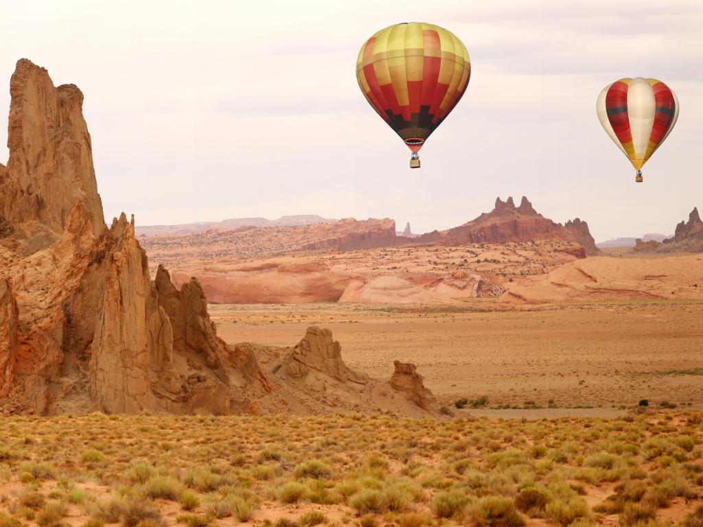 Colorful hot air balloons fly across arid desert landscape with jagged rock formations rising up