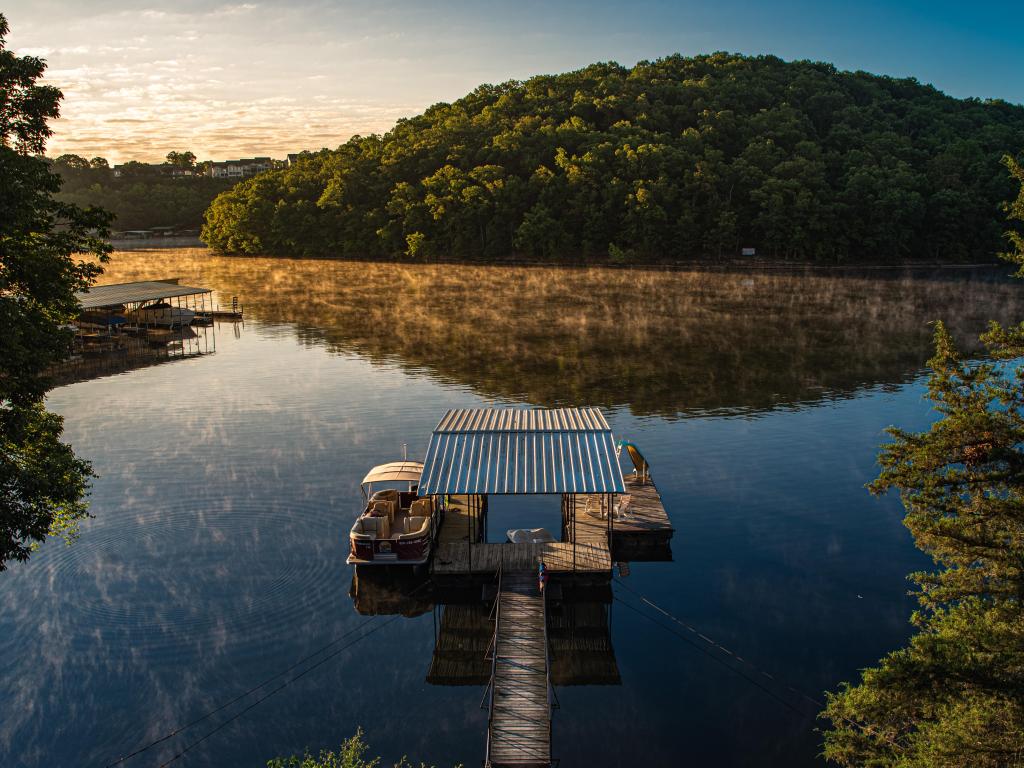 Osage Beach, Missouri, USA with early morning sunlight illuminated wood covered hills across a calm reflecting lake with dock in foreground.