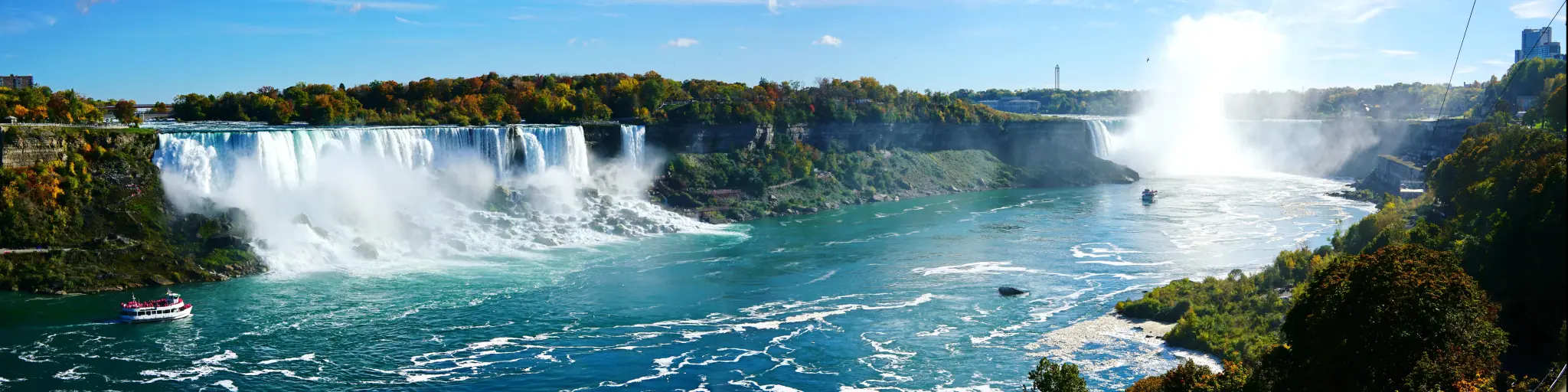 Niagara Falls, USA/Canada taken as a panoramic shot of the waterfalls on a sunny day with blue sky and boats in the water in the foreground. 