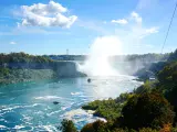 Niagara Falls, USA/Canada taken as a panoramic shot of the waterfalls on a sunny day with blue sky and boats in the water in the foreground. 