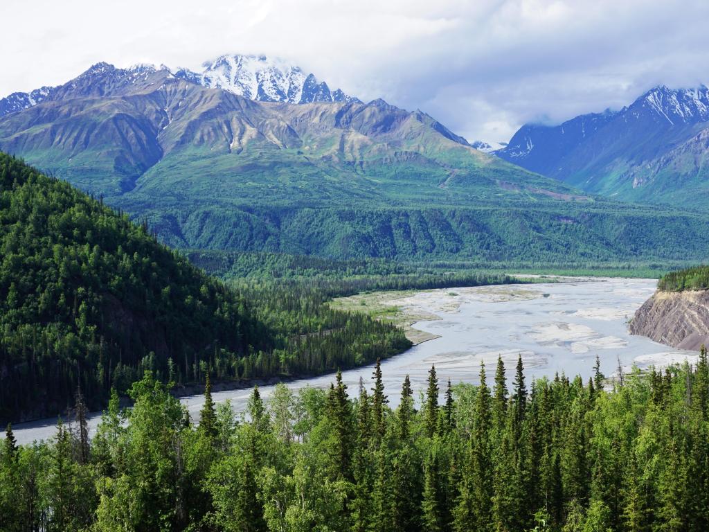 The Susitna River and Valley in Alaska with trees lining the mountains and snow-capped mountains in the distance under a cloudy sky.