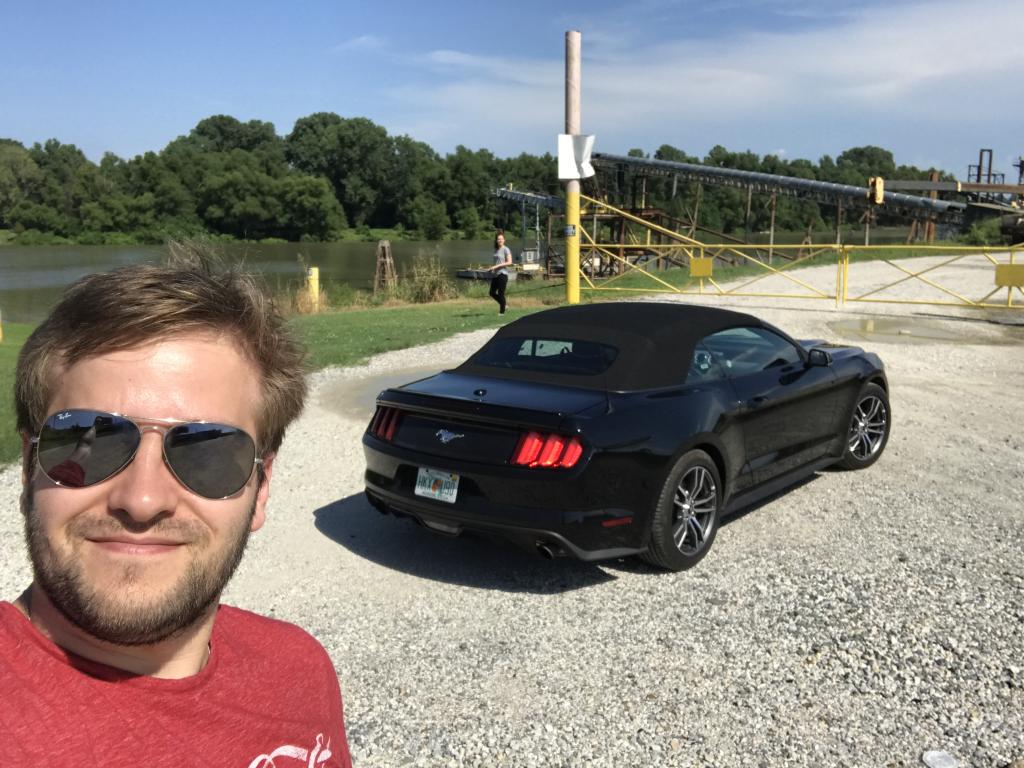 Sasha taking a selfie next to a black Mustang convertible by the Mississippi River.