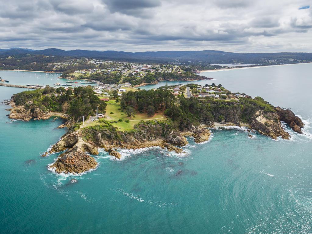 Eden, NSW, Australia showing an aerial shot of the dramatic edges of land surrounded by a turquoise sea and the town beyond in the background.