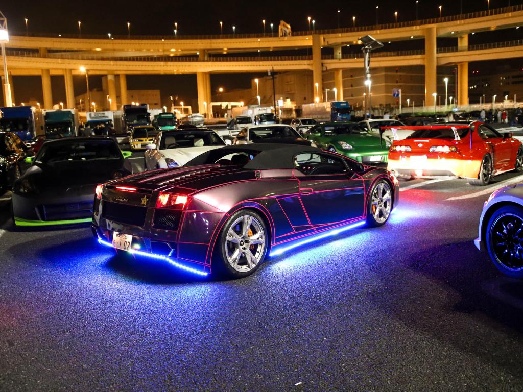 A car meeting at Daikoku Futo in Tokyo with a dark car in focus at night time. It is illuminated by blue lights underneath.