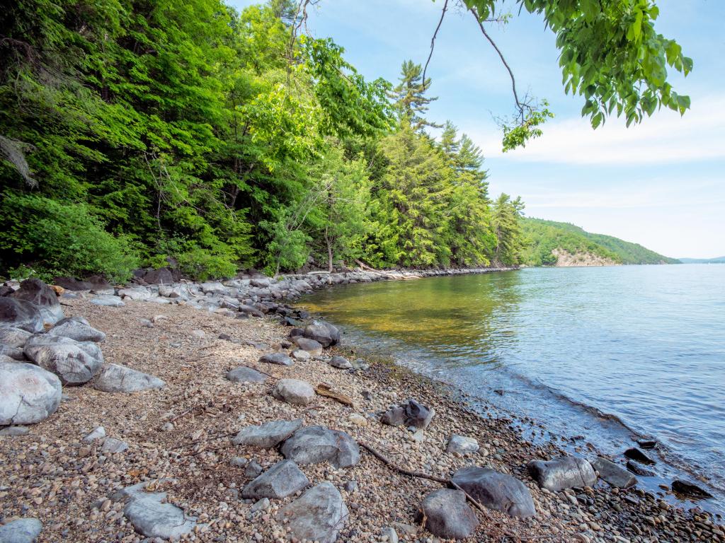 Lake Champlain, USA/Canada border taken at a bay with a rocky shore and tall trees in the distance on a sunny day.