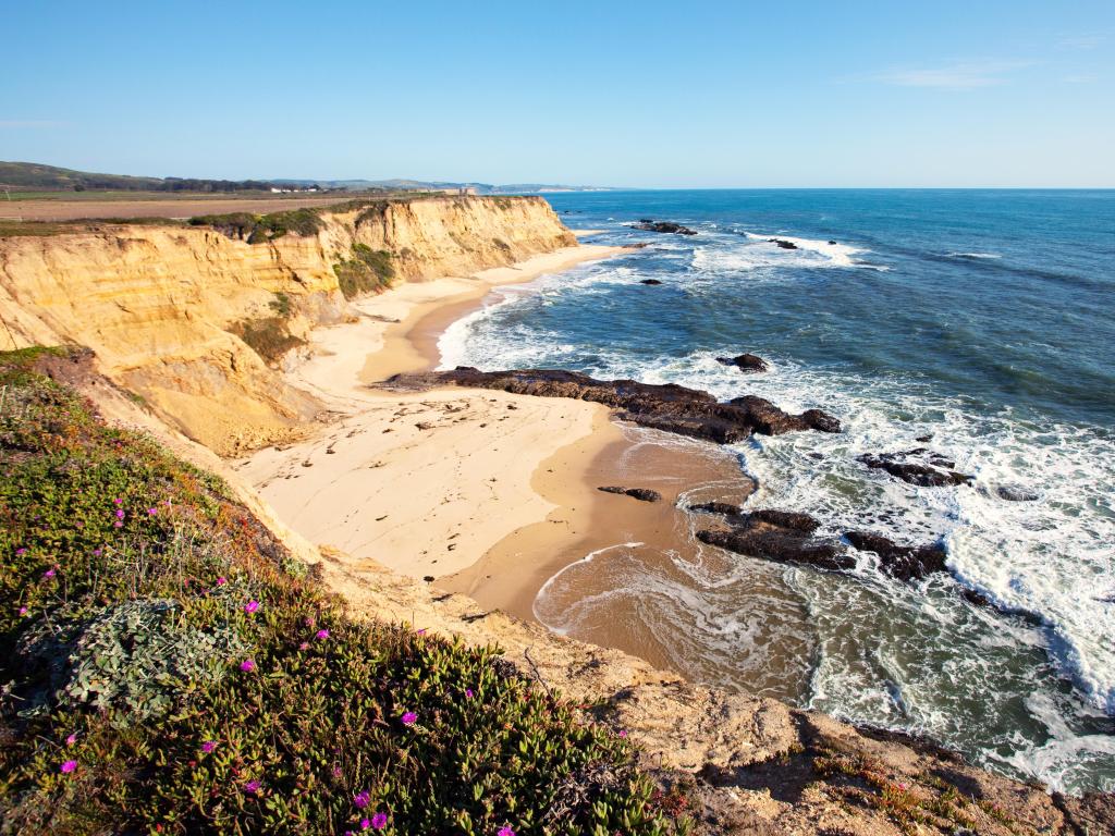 Steep sandy cliffs and sandy beach in a wide bay with deep blue ocean and waves breaking on rocks