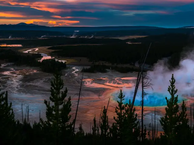 Yellowstone National Park, USA taken after sunset at the Grand Prizmatic Overlook with trees in the foreground and the steam from the craters in the distance.