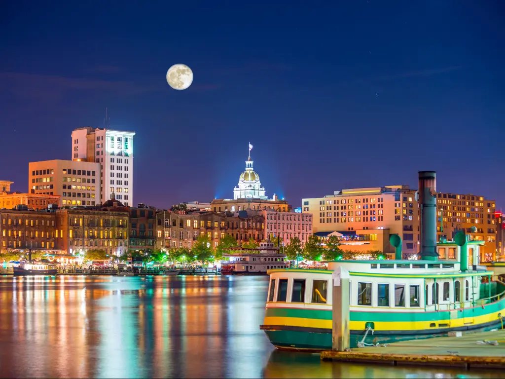 Historic waterfront buildings seen across river at night with river boat in foreground and moon