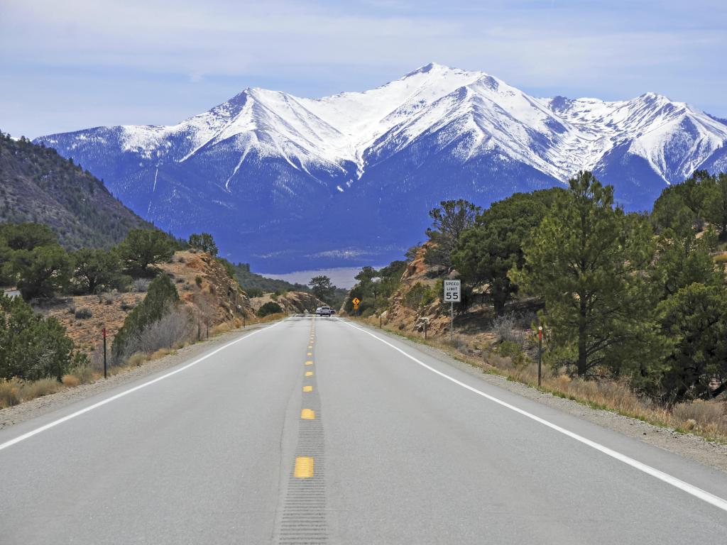 Straight road with green trees either side leads towards rugged, snow-capped mountains
