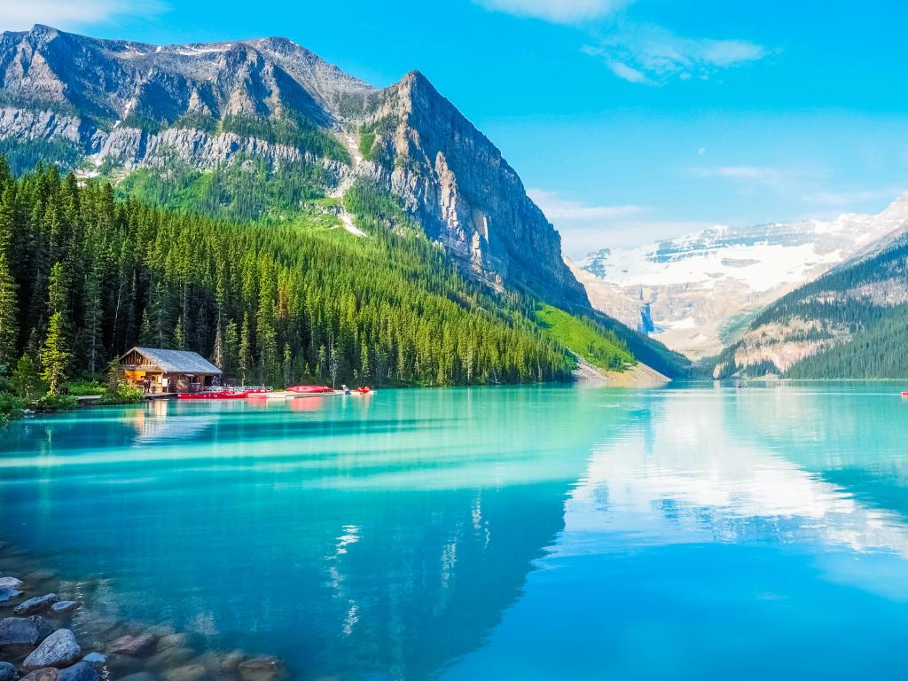 Banff National Park, Canada with stunning turquoise Lake Louise in the foreground, tall trees in the shore beyond and huge mountains in the distance on a bright sunny day.