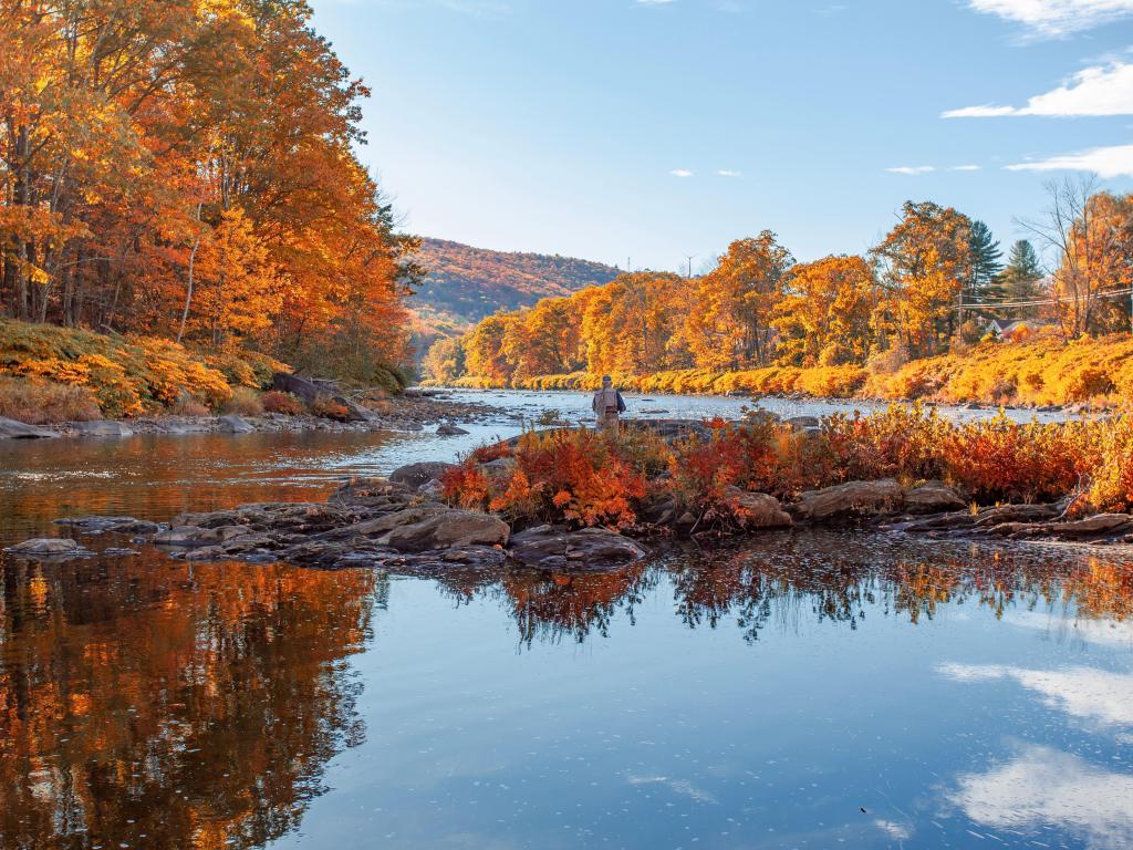 Fisherman stands by calm river in a valley showing vibrant orange and red fall foliage
