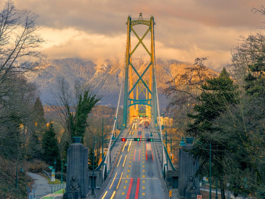 Golden sunset light illuminates a tall bridge with parkland to either side and a cloud-covered mountain on the far side of the river