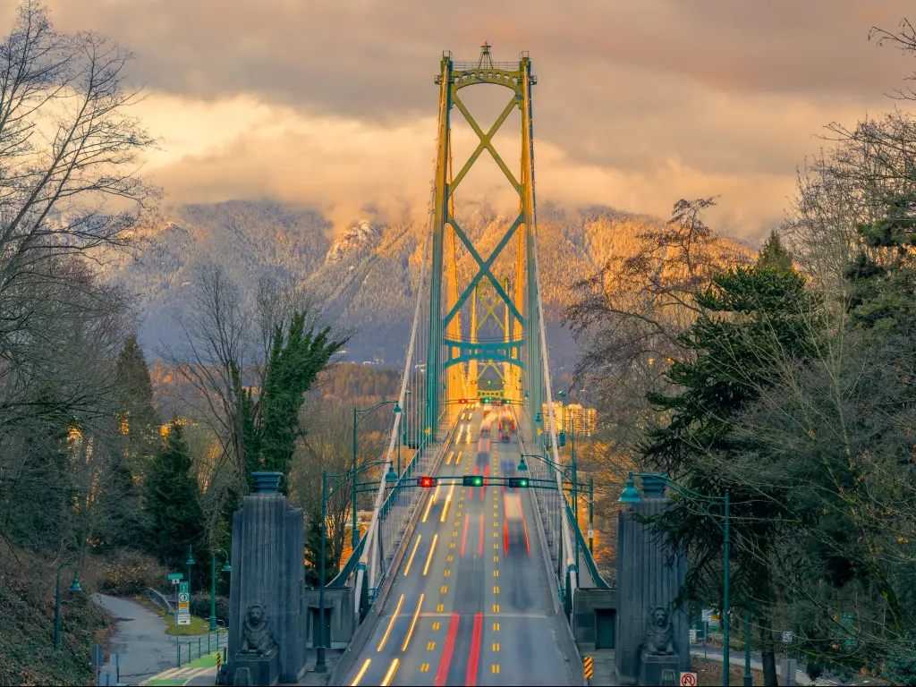 Golden sunset light illuminates a tall bridge with parkland to either side and a cloud-covered mountain on the far side of the river