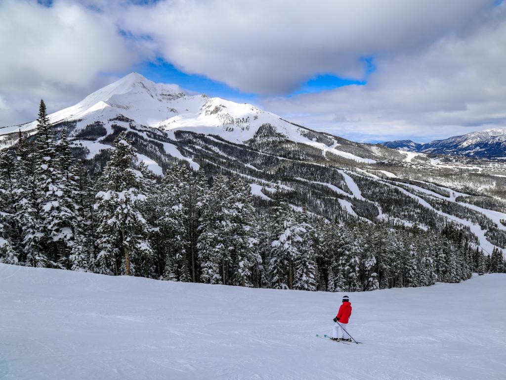 Big Sky, Montana, USA with a man in red downhill skiing and the snow-capped mountain in the distance taken on a sunny but cloudy day.