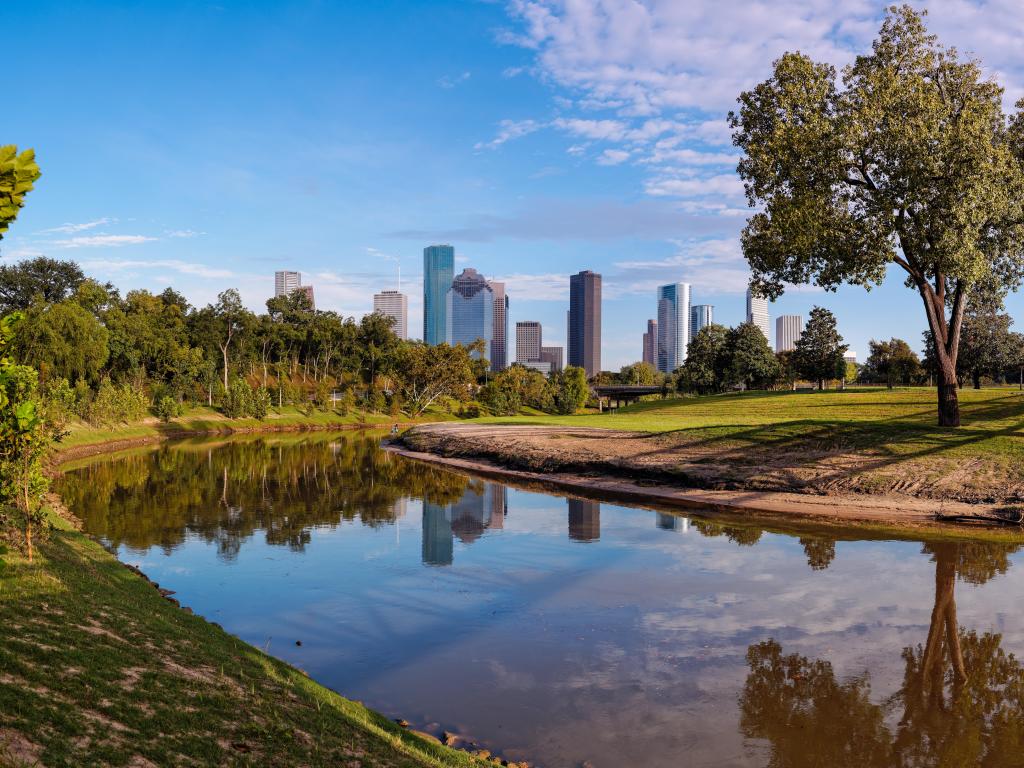Buffalo Bayou Park, USA with a large lake in the foreground surrounded by trees and grass and the downtown skyline in the background on a sunny day. 