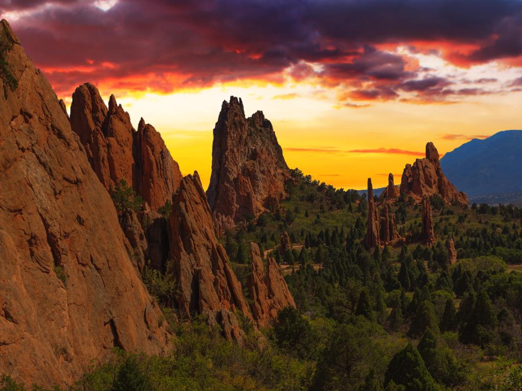 Deep orange rock formations stand up out of green forest with a dark red sky