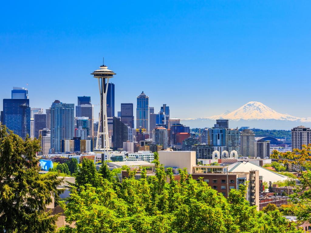 Seattle, Washington with the downtown skyline in the background and Mt. Rainier in the distance, and green trees in the foreground under a blue sky.