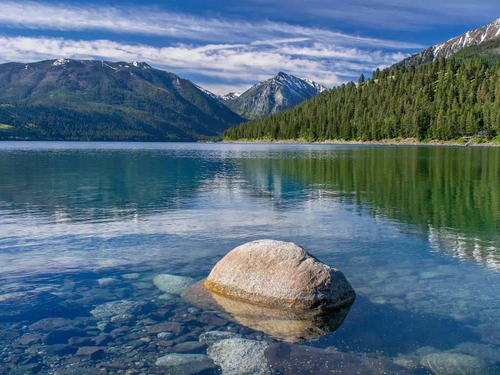 Wallowa Lake glistening in the sunshine, with trees around the shore and a rock reflecting in the water's surface