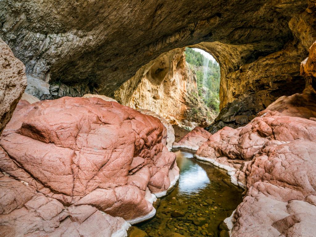 Tonto Natural Bridge State Park, Arizona, USA with a stream in the foreground surrounded by rocks and leading to a rocky arch in the distance.