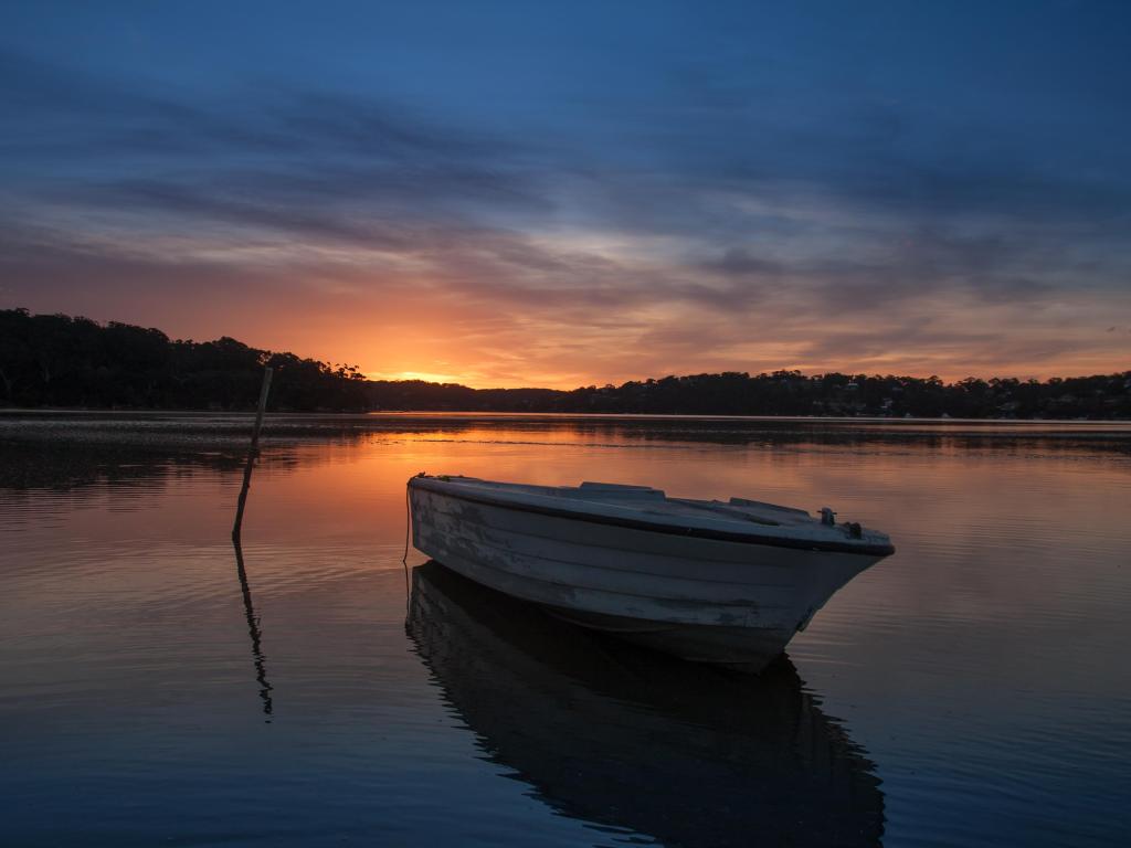 Small boat moored on lake at sunset with pink sunlight reflected on the water and trees shown in silhouette 