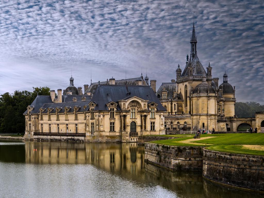 Panoramic view of the Château de Chantilly reflected in the water of the river with light clouds in the sky.