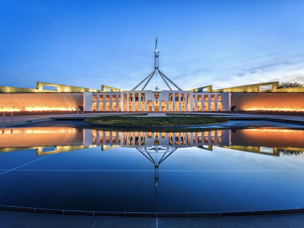 Wide modern white building with tall, narrow windows in central section, with deep blue sky behind. Buildings and sunset reflected into still water in front